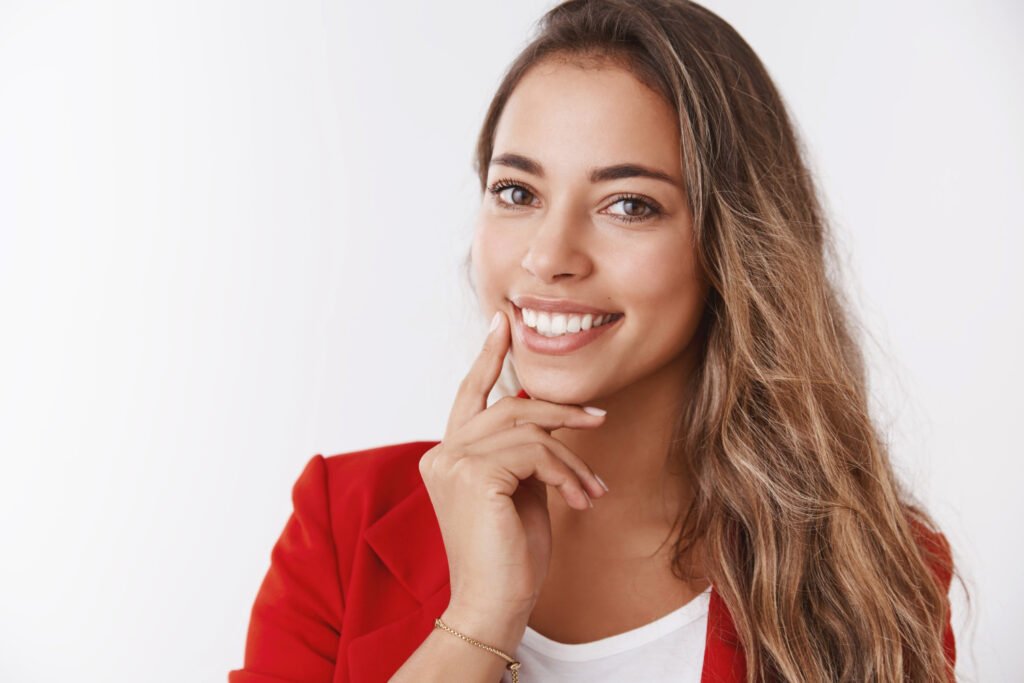 Mulher de blazer vermelho toca o queixo sorrindo. Saiba os riscos da cirurgia ortognática na Dra. Viviane Amaral em Brasília - Asa Sul.